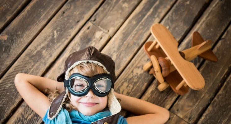 Child in aviator cap and goggles lying beside a toy wooden airplane